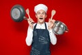 Shocked chef in uniform holds many kitchen items on red isolated background, kitchen worker in stress