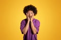 Shocked confused adult african american curly man in purple t-shirt with open mouth presses hands to face