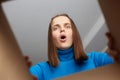 Shocked disappointed woman customer wearing blue turtleneck opening cardboard box with broken or wrong order, looking inside