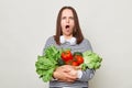 Shocked displeased brown haired young woman embraces bouquet of fresh vegetables wearing striped casual shirt isolated over gray Royalty Free Stock Photo
