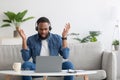 Shocked displeased young black bearded man in headphones raises hands, talk by laptop and gesturing