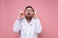 Shocked doctor in a white coat and glasses points index fingers up and looks frightened. Pink background.