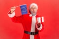Shocked elderly man with gray beard wearing santa claus costume with open mouth waving Europe flag and holding in hands wrapped