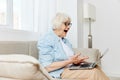 a shocked elderly woman is sitting on a comfortable sofa holding a laptop on her lap and looking very scared at the Royalty Free Stock Photo