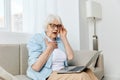 a shocked elderly woman is sitting on a comfortable sofa holding a laptop on her lap and looking very scared at the Royalty Free Stock Photo