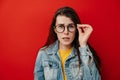 Shocked emotional young woman keeps hand on rim of spectacles, dressed in denim jacket, gazes with stupefaction Royalty Free Stock Photo