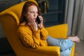 Shocked excited young woman talking on smartphone sitting in yellow chair at home. Pretty puzzled redhead female having Royalty Free Stock Photo