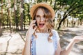 Shocked fair-haired girl standing in park and talking on phone. Outdoor shot of beautiful young wom Royalty Free Stock Photo