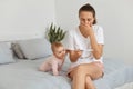 Shocked female with pigtail holding pregnancy test in hands, sitting on bed in bedroom with her infant daughter, covering mouth