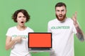 Shocked friends couple in volunteer t-shirt isolated on green background. Voluntary free work assistance help charity