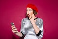 Shocked ginger woman holding cheek while using smartphone