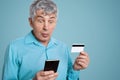 Shocked grey haired male looks with frustrated expression at smart phone, can`t understand how use mobile application and do onlin