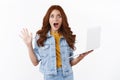 Shocked and impressed cute redhead female student in denim jacket, hold laptop, raise hand up in triumph and celebration