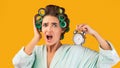 Shocked Lady With Curlers Holding Alarm Clock On Yellow Background