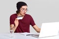 Shocked male with appealing appearance, holds plastic card in hand, being terrified as checks bank account on laptop computer,