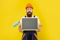 shocked man renovator in helmet and work clothes hold paint roller brush and blackboard