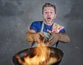 Shocked messy man with apron holding pan in fire burning the food in kitchen disaster and unskilled and unexperienced terrible ho