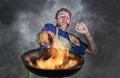 Shocked messy man with apron holding pan in fire burning the food in kitchen disaster and unskilled and unexperienced terrible ho