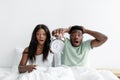 Shocked millennial black guy and woman wake up after night with open mouths on bed in bedroom, lady hold alarm clock