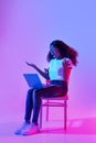 Shocked millennial black woman using laptop, opening mouth in surprise, sitting on chair in neon light, copy space