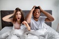 Shocked multiracial couple with alarm clock being late to work, sitting on bed, grabbing their heads in terror
