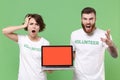 Shocked nervous friends couple in volunteer t-shirt isolated on green background. Voluntary free work assistance help