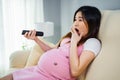 Shocked pregnant woman watching tv with remote control on sofa in living room Royalty Free Stock Photo