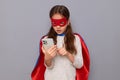 Shocked sad little girl wearing superhero costume and mask isolated over gray background using cell phone for online communicating Royalty Free Stock Photo