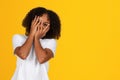 Shocked scared shy teenage black girl in white t-shirt covers face with hands, isolated on yellow background Royalty Free Stock Photo