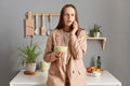 Shocked scared woman with brown hair wearing beige jacket standing in home kitchen interior talking on smart phone while having Royalty Free Stock Photo