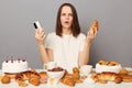 Shocked scared woman wearing white T-shirt isolated over gray background sitting at table with desserts holding mobile phone and Royalty Free Stock Photo