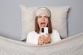 Shocked scared young woman wearing white T-shirt and sleep eye mask lies under blanket in bedroom isolated on gray background Royalty Free Stock Photo