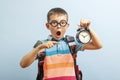 Shocked schoolboy in glasses with backpack holds an alarm clock in his hand and points at it with his finger, on blue background. Royalty Free Stock Photo