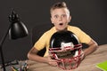 shocked schoolboy sitting with american football helmet at table with eyeglasses lamp colour pencils and books on grey