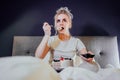 Shocked, stressed, surprised woman eating cake and holding a remote control while watching TV in bed. Scary horror movie Royalty Free Stock Photo