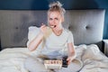 Shocked, stressed, surprised woman eating ice cream, holding a remote control while watching TV in bed. Scary horror Royalty Free Stock Photo