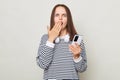 Shocked surprised brown haired woman wearing striped shirt posing over gray background holding smart phone covering mouth