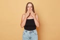 Shocked surprised brown haired young woman wearing black top and jeans standing isolated over beige background looking at