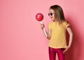 Amazed blonde kid girl in yellow t-shirt, shiny pink leggings and sunglasses looks at big pink lollipop she holds