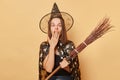 Shocked surprised young woman wizard wearing witch costume holding in hand broom  over beige background amazed halloween Royalty Free Stock Photo