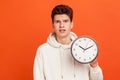Shocked teenager in casual white hoodie afraid of being late, holding in hand wall watch, deadline, punctuality