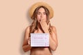 Shocked terrified female covers mouth with palm, shcoked to have periods, holds period calendar, wears bikini and straw hat, isola