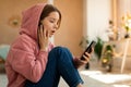 Shocked upset teen girl reading bad news message on cellphone, touching cheek in amazement, sitting at home