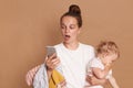 Shocked woman with bun hairstyle wearing white t shirt standing with baby kid and using cell phone, reading surprised news,