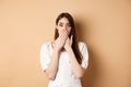 Shocked woman covering mouth with hands and staring startled at camera, standing speechless from gossips on beige Royalty Free Stock Photo