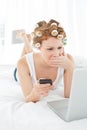 Shocked woman in hair curlers with cellphone looks at laptop in bed Royalty Free Stock Photo