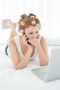 Shocked woman in hair curlers using cellphone and laptop in bed Royalty Free Stock Photo