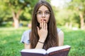 Shocked woman sitting in park while holding book and looking at the camera Royalty Free Stock Photo