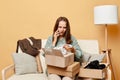 Shocked woman sitting on sofa among boxes with clothing against beige wall opening parcel from internet shore talking phone having