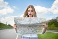Shocked young blonde woman hitchhiking on road, checking map, feeling lost and confused, traveling alone by autostop